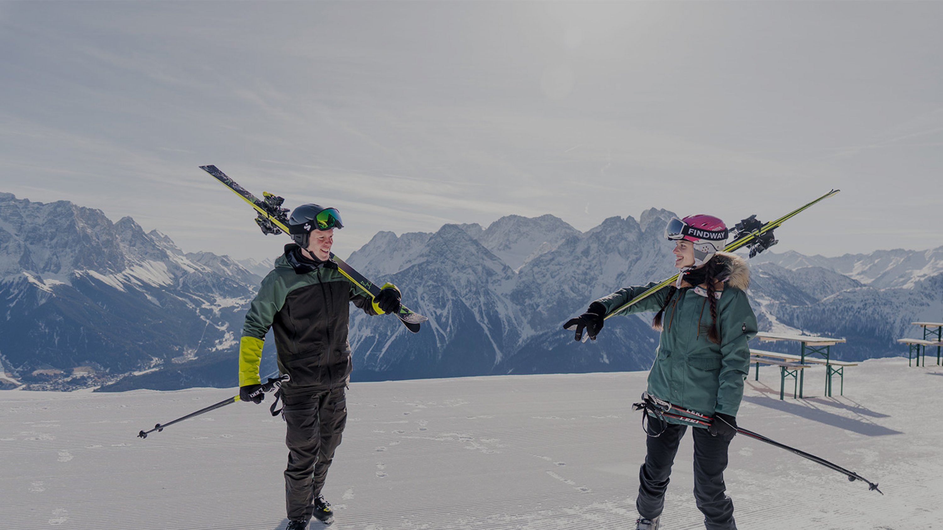 Zwei Skifahrer tragen Ihre Ski auf den Schultern, im Hintergrund ein winterliches Bergpanorama
