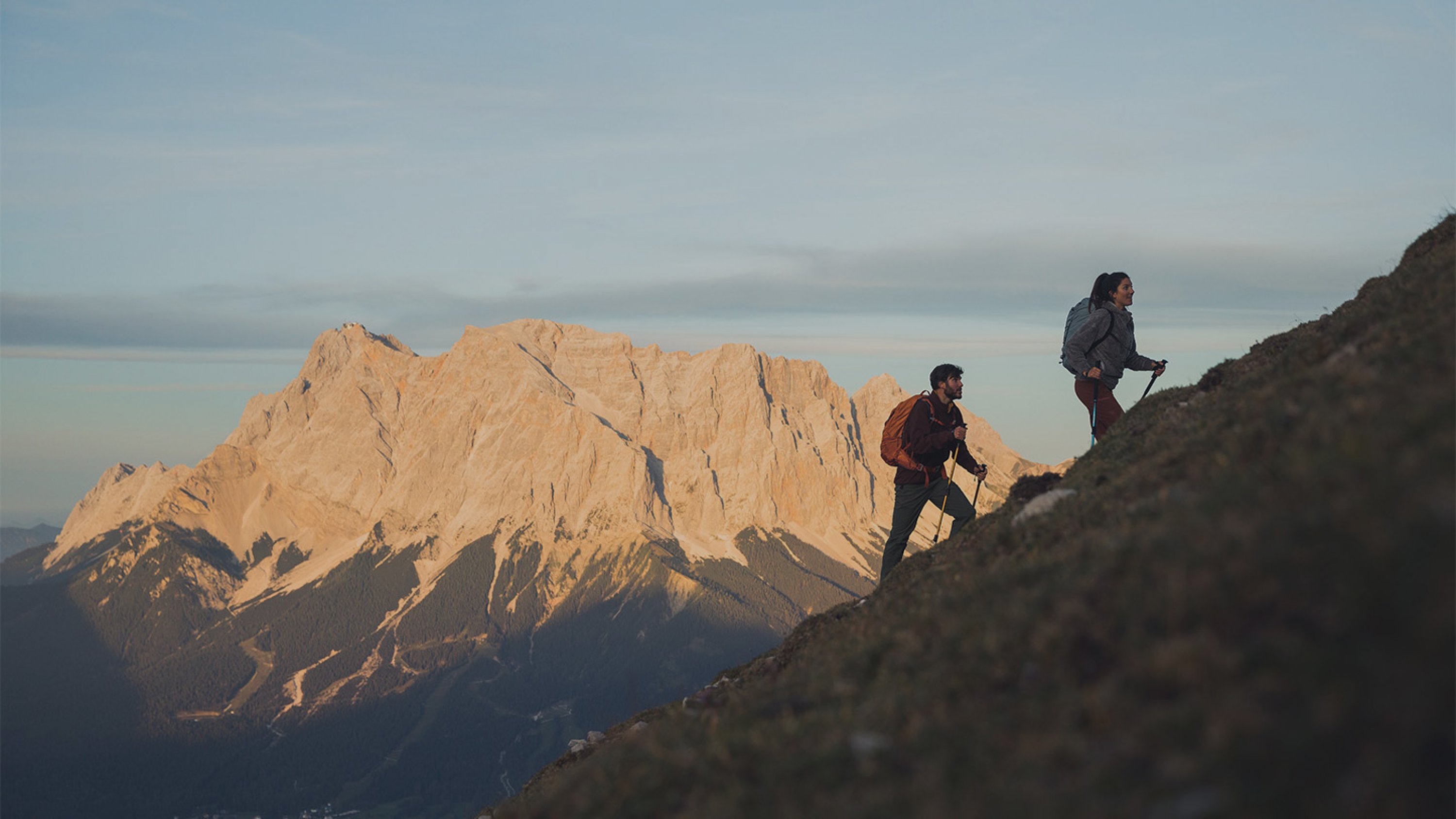 A woman and a man hike up a steep mountain hillside, in the background a large mountain massif illuminated by the sun