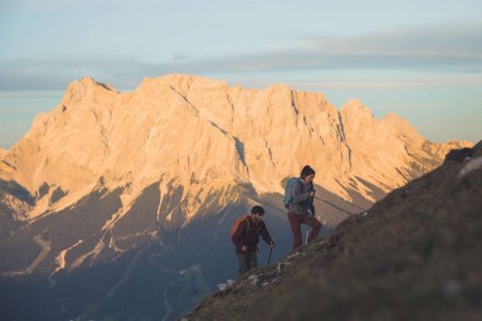 Eine Frau und ein Mann wandern einen steilen Berghang hinauf, im Hintergrund ein großes Bergmassiv, welches von der Sonne angestrahlt wird | © Tiroler Zugspitz Arena/C. Jorda