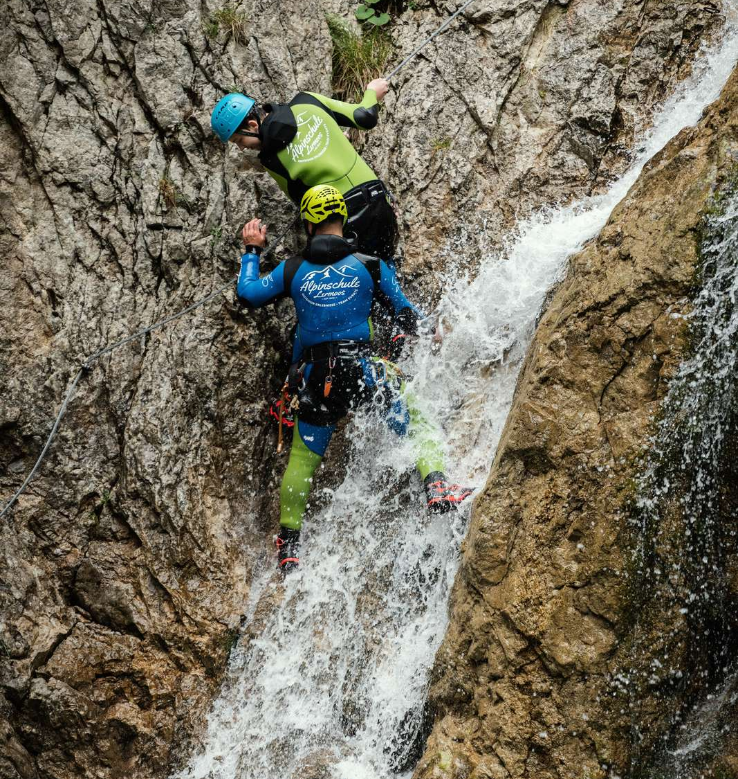 Two people are doing canyoning, they climb down a waterfall while holding on to a rope.