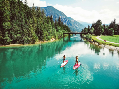 Zwei Stand-Up-Paddler auf dem türkisen Wasser des Heiterwanger Sees, im Hintergrund eine Brücke und Bergpanorama | © TZA