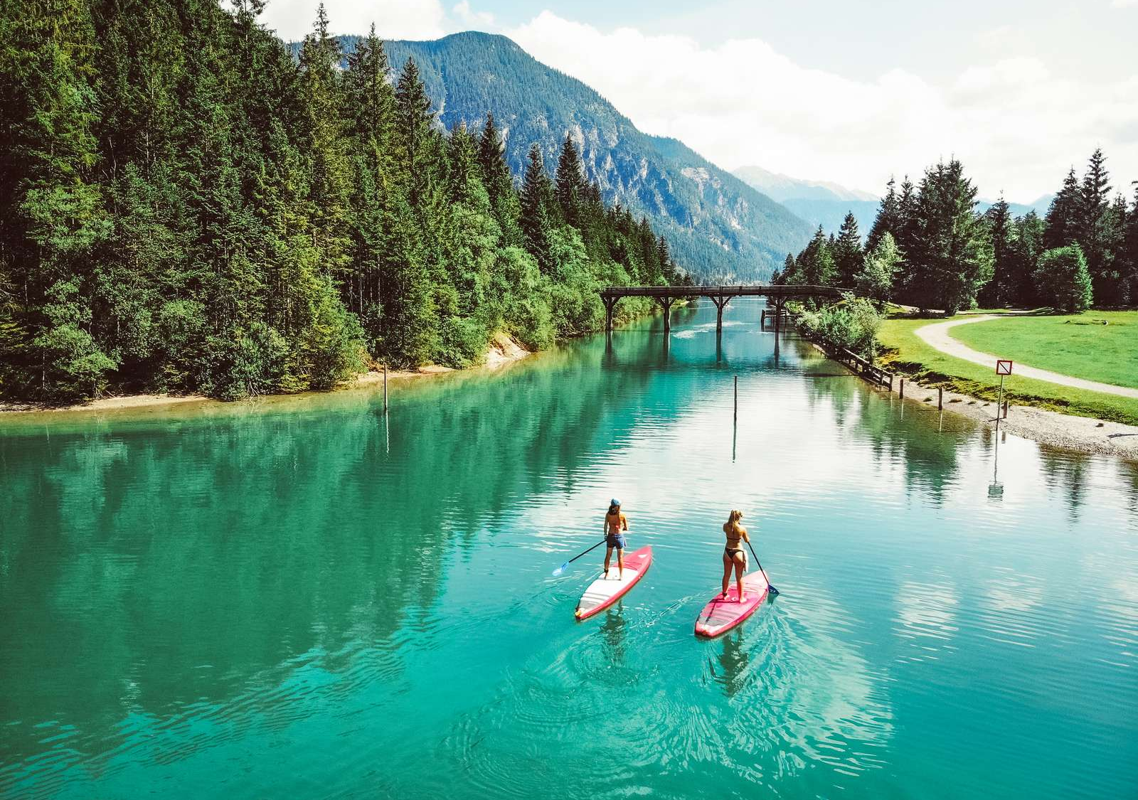 Two stand-up paddlers on the turquoise waters of Lake Heiterwanger See, with a bridge and mountain panorama in the background