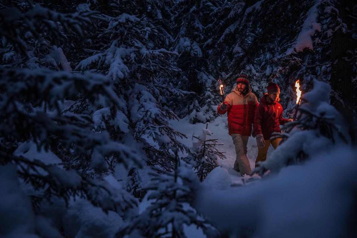A couple walks through a snowy forest at night with torches